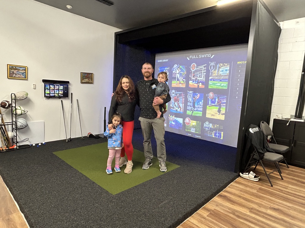 Apex-Play-Coach-Mike-family A man and a woman stand on an indoor golf simulator mat with two small children, one held by the man and the other standing beside the woman. Golf equipment and chairs are visible in the background.
