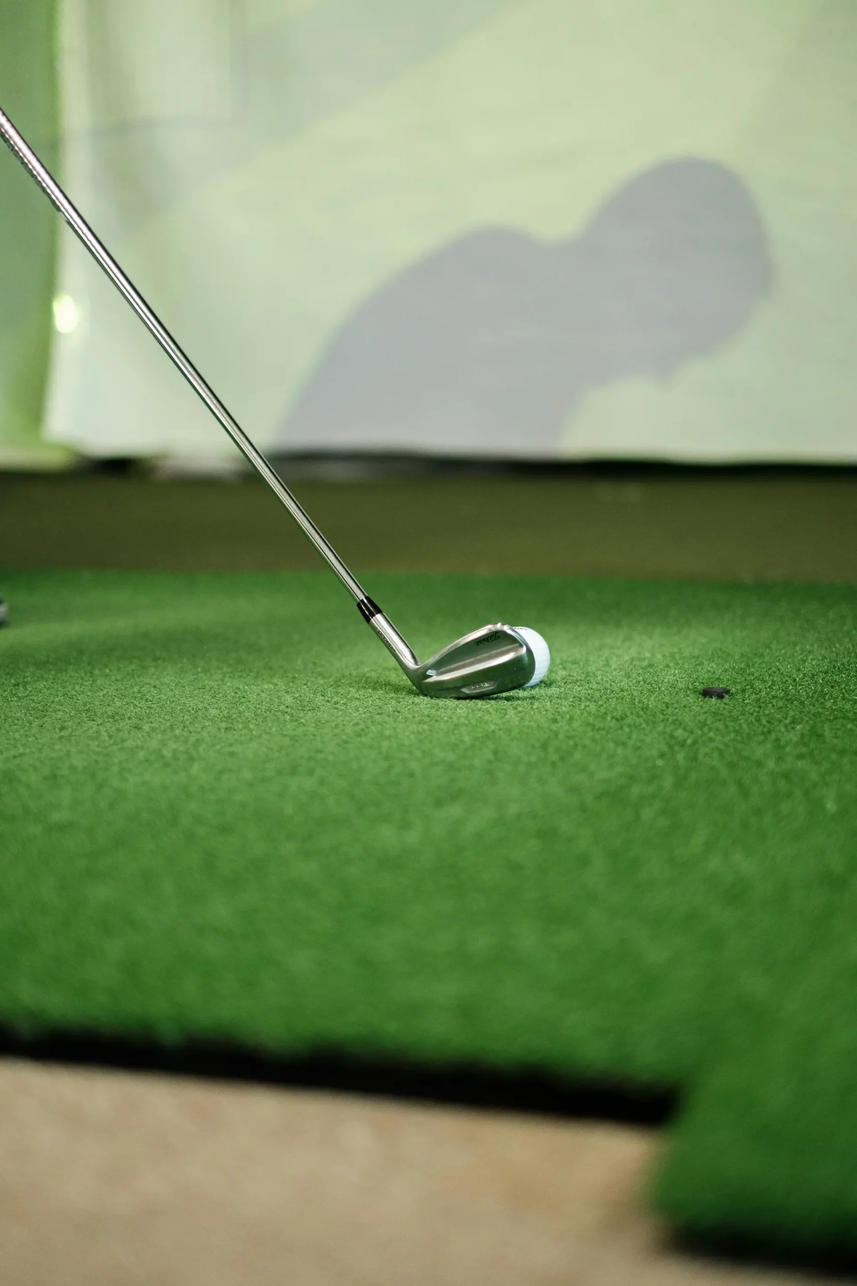 golf-swing-indoors A close-up of a golf club positioned behind a golf ball on green artificial turf, preparing for a putt. The background is blurred, with a shadow visible on the wall.