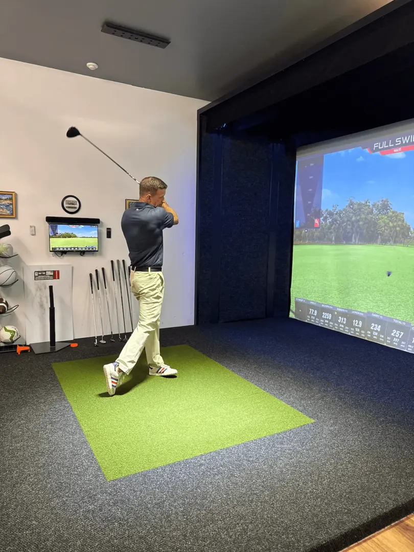 Driver-on-golf-simulator-Apex-Play A person in light pants and a dark shirt swings a golf club on an indoor simulator, with a large screen displaying a virtual golf course and golf equipment visible on the wall and floor nearby.