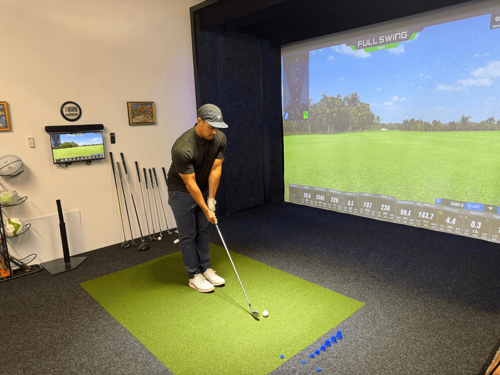 Full-swing-tee-set-up-Apex-Play A person wearing a cap and dark clothing prepares to hit a golf ball with a club indoors, facing a golf simulator screen displaying a virtual golf course. Several clubs and golf balls are arranged nearby.