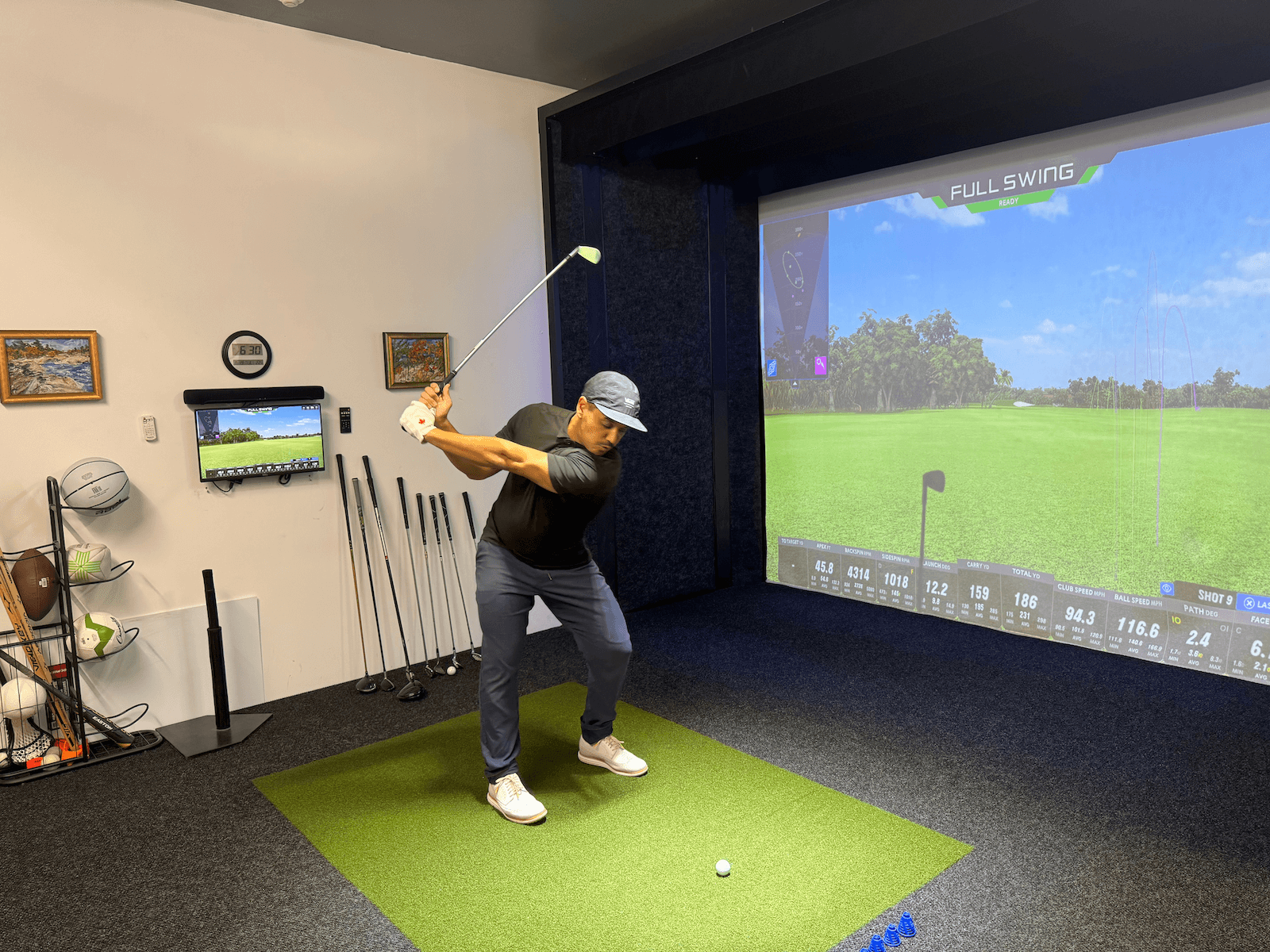 Man-at-the-apex-of-his-golf-swing-apex-play (1) A man stands on artificial turf indoors, preparing to swing a golf club in front of a large golf simulator screen displaying a virtual golf course. Golf clubs, balls, and equipment are visible nearby.