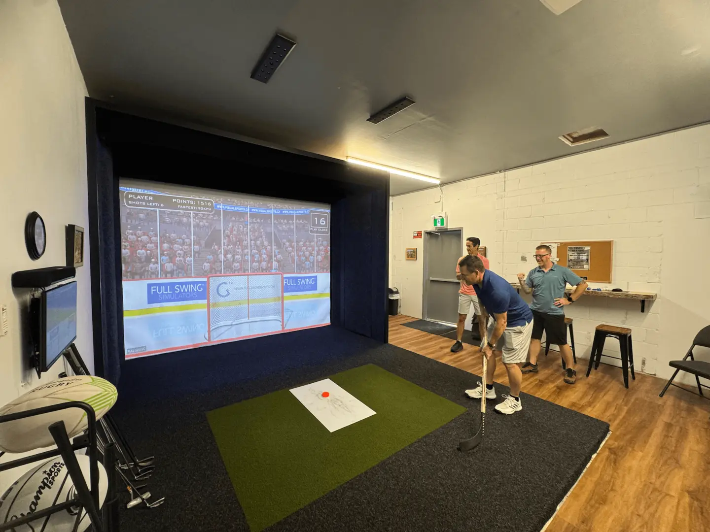 Man-prepares-to-hit-a-hockey-puck-on-a-simulator-apex-play-1 Three men stand in a room with wood floors and white walls, using a golf simulator. One man is swinging a golf club at a ball on artificial turf while the others watch. A virtual golf course is displayed on a large screen.