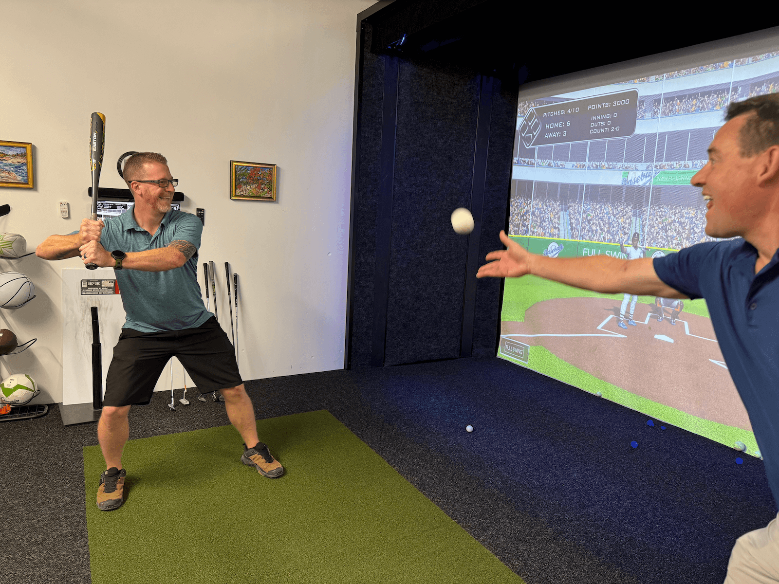 Man-throws-a-ball-to-someone-readdy-to-hit-it-on-a-simulator-apex-play (1) Two men play a virtual baseball game indoors; one is pitching a ball while the other holds a bat, ready to swing. A sports simulator screen displays a baseball stadium in the background. Sports equipment is visible on the left.