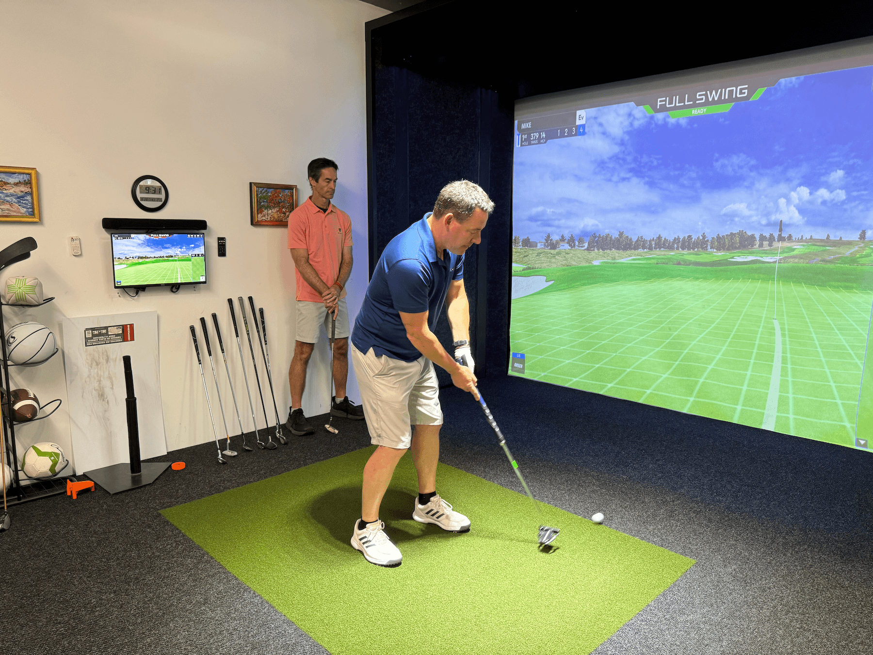 A man in a blue shirt and white shorts prepares to hit a golf ball in an indoor golf simulator, while another man in a pink shirt stands nearby. Golf clubs and a monitor are visible on the left side.