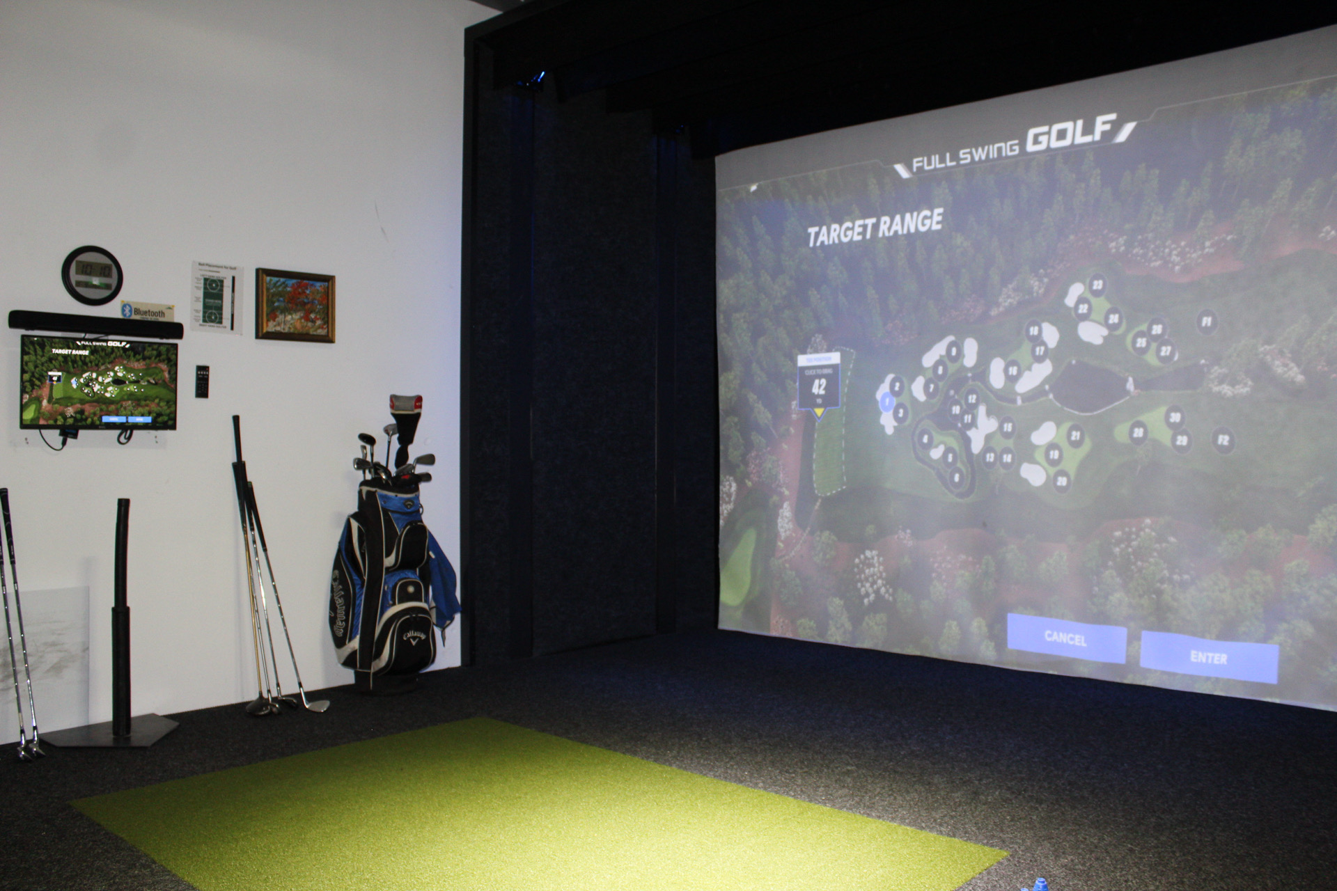 3DE2F3D7-8A87-440D-89EF-D6696C451F68 A golf simulator setup with a large projection screen displaying a virtual golf course, a green artificial turf mat, several golf clubs in a bag, and a wall-mounted TV showing a golf game.