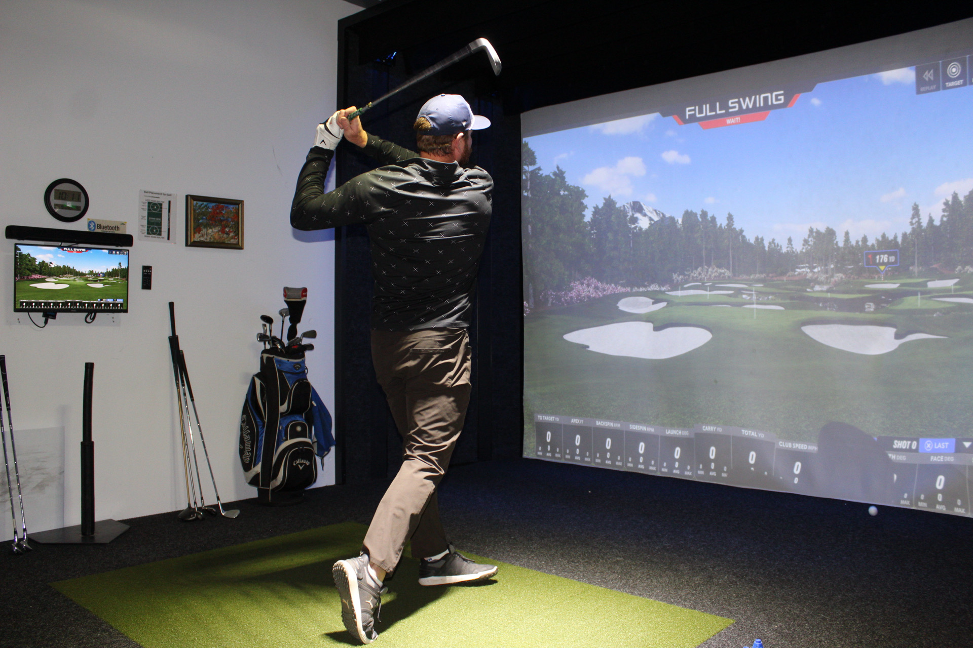 5D5D48B9-FA58-4644-BDDE-2DF4E61F1E0F A person swings a golf club indoors on a simulator, aiming at a virtual golf course projected on a large screen. Nearby are golf clubs, a guide monitor, and a green putting mat.