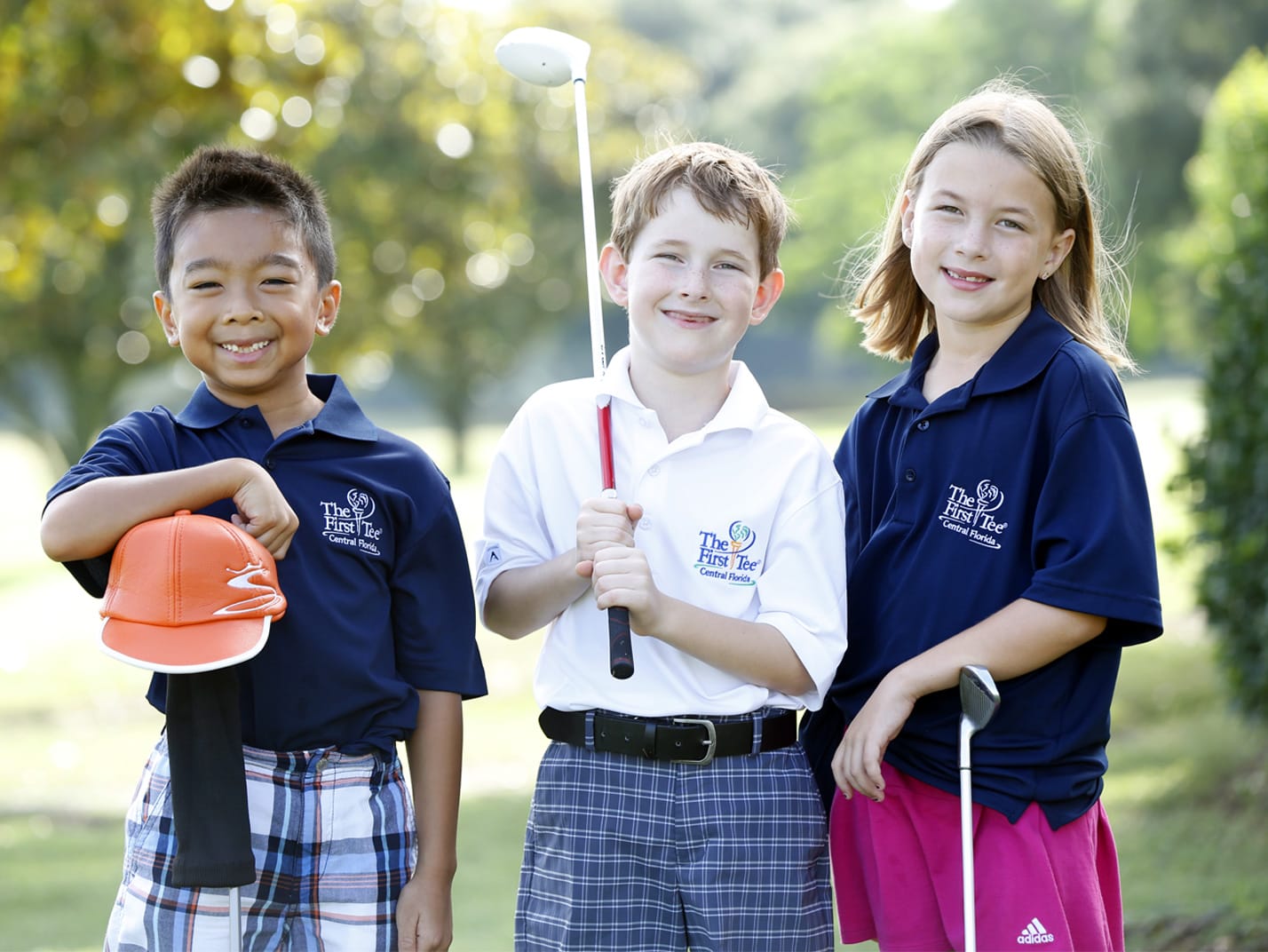 Three children stand outdoors holding golf clubs and smiling. They wear “The First Tee” shirts and are dressed in golf attire. Trees and greenery are visible in the background under bright daylight.